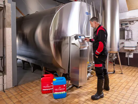 A man and boy stand in a dairy facility. The man drinks a glass of milk while leaning on a Lely cooling tank for the Lely Astronaut, and the boy smiles at him.