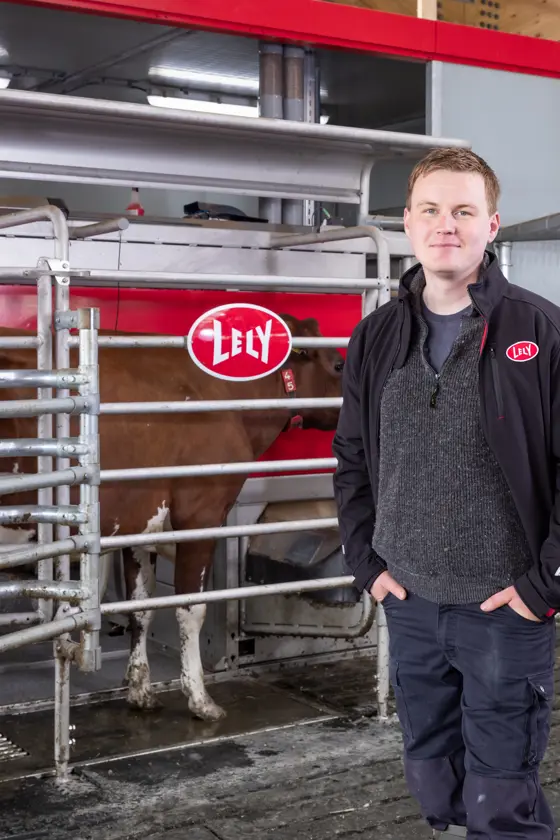 A man in a black shirt and cap stands in front of a Lely automated milking system in a barn, with a cow inside the machine behind him.